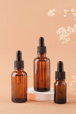 Beyaz podyumun üzerinde siyah kapaklı kehribar cam damlacıklar bej arka planda kuru bitkilerle ürün sunumu için