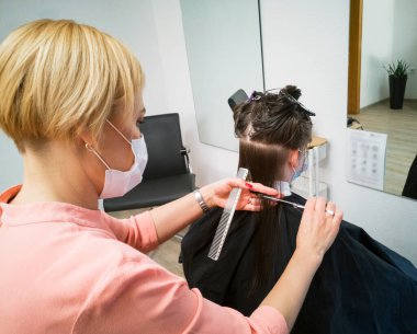 Siyah-19 güvenlik önlemleri için tıbbi maskeli sarışın kuaför esmer kadın müşteri saçını kesti. Güzellik salonunun yeniden açılması, pandemik karantina. Klasik kuaför teknolojisi..