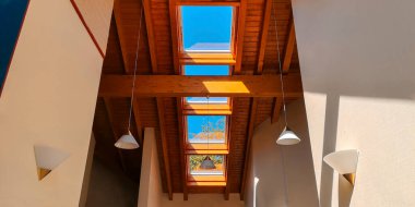 View of ceiling of gable roof without attic with three plastic windows, finished with clapboard,wooden beam,identical suspended white frosted lamps,2 symmetrically on white walls.Selective focus.