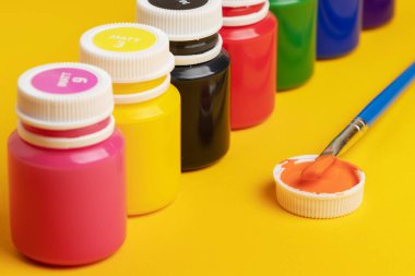 Sarı arka planda akrilik boya ve fırçayla dolu kavanozlar.