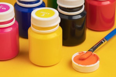 Sarı arka planda akrilik boya ve fırçayla dolu kavanozlar.