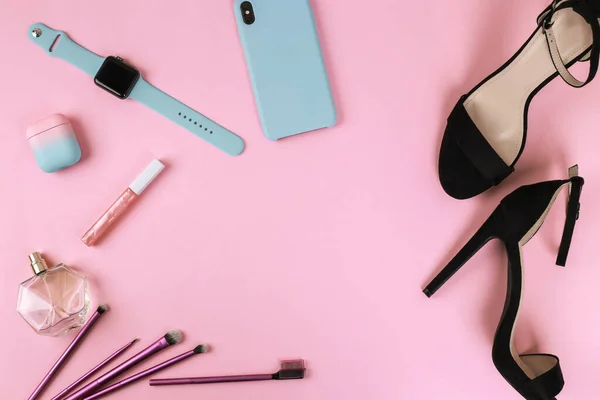 Smart gadgets on pink background. Phone, headphones, smart watch and lip gloss, perfume, black sandals and makeup brushes. Top view with copy space, flat lay. Woman devices
