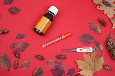 Concept of health care, baby treatment and medicine - a bottle of medicines or antipyretic syrup and a measuring syringe, thermometer on a red background with leaves. Diseases in the fall clipart