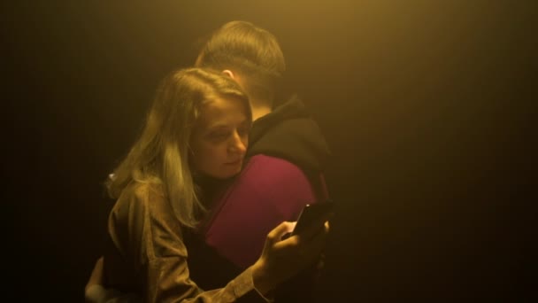 Pasangan cantik dalam cinta Kaukasia pria dan gadis berdiri merangkul dan melihat smartphone pada latar belakang hitam. Sandera teknologi baru dan smartphone, gerakan lambat — Stok Video