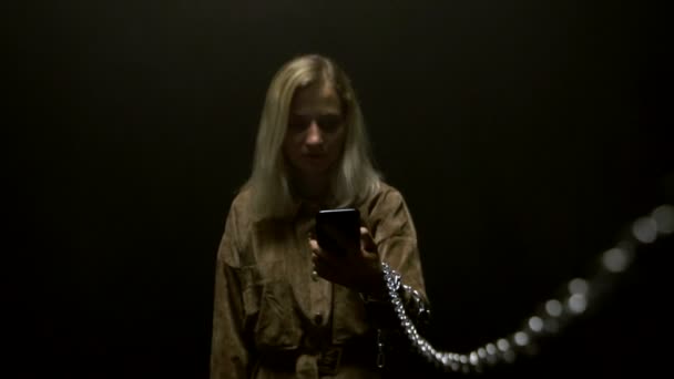 Una joven caucásica sostiene un teléfono inteligente en su mano y lo mira, una cadena de metal está atada a su mano. Concepto de rehenes de nuevas tecnologías y redes sociales, fondo negro, cámara lenta — Vídeo de stock