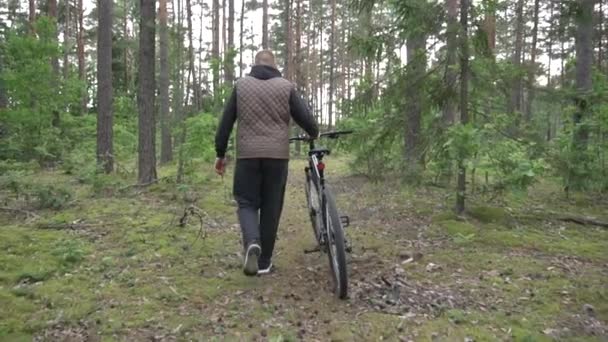 En mann i hette og vest ruller en sykkel gjennom skogen. Sykkel- og reisekonsept, langsom bevegelse – stockvideo