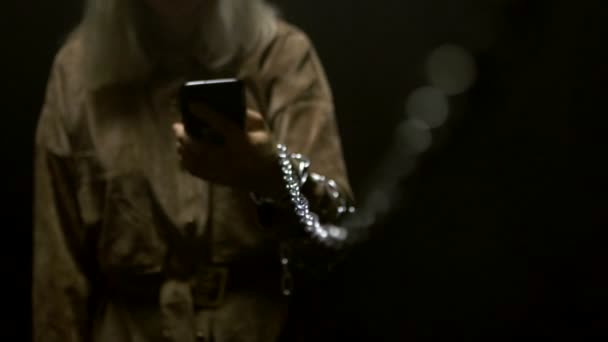 Ein junges kaukasisches Mädchen hält ein Smartphone in der Hand und schaut hinein, eine Metallkette ist an ihrer Hand gebunden. Geiselnahme-Konzept neuer Technologien und sozialer Netzwerke, schwarzer Hintergrund, Zeitlupe — Stockvideo