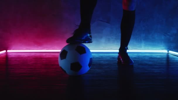 サッカー選手は足で床にボールを転がす。サッカートレーニングの概念。マゼンタブルーのライトの暗いスタジオでボールを持つアスリートスポーツマン。スポーツだ。Blackmagic ProResで撮影。ベット — ストック動画
