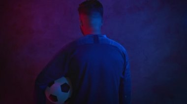 Futbol konsepti. Koltukaltında top olan oyuncu poz veriyor, arkasını dönüp karanlık bir stüdyoda kameraya bakıyor. Spor. Portre. ProRes 'ta Kara Büyü Çekimi