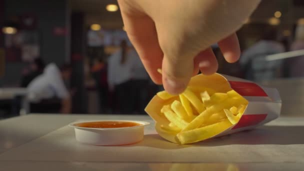 Frietjes liggen op tafel in café restaurant, met de hand neemt en doopt aardappelen in saus ketchup. Ongezonde levensstijl, calorierijke fastfood. Bezoekers op de achtergrond maken orde in de buurt van checkout — Stockvideo