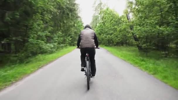 Guy slowly rides a cross-country mountain bike on an asphalt road through the green forest. View of the cyclists back. Sport, recreation and pastimes, health benefits, fitness. Trip journey — Stock Video
