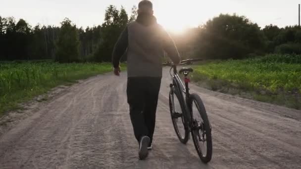 Man walks and rolls a bicycle next to him along a dirt sand road at sunset. Bike malfunction, damage, breakdown. View of the cyclists back. Sport, recreation, health benefits, fitness. Trip journey — Stock Video