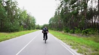 Sırt çantalı bir adam yeşil ormanda asfalt yolda hızla dağ bisikletine biniyor. Bisikletçilerin geri dönüşünü izle. Spor, eğlence ve hobi, sağlık sigortası, fitness. Yolculuk