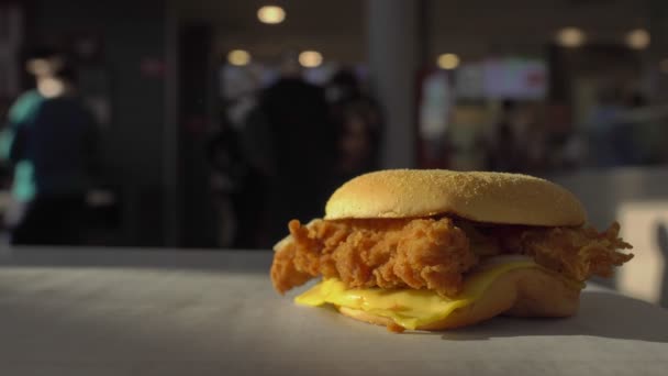 Fast food burger z kotletem kurczaka leżą na stole wewnątrz kawiarni restauracji. szkodliwe jedzenie. Niezdrowy styl życia, smażone i wysokokaloryczne. Goście w tle złożyć zamówienie w pobliżu kasy. Żywność szybka. — Wideo stockowe