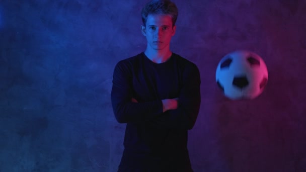 Fotbollsspelare poserar i en mörk studio. Fotboll och sport koncept. Ung kaukasisk idrottsman tittar på kameran, en boll studsar i närheten. Porträtt. Skjuten på Blackmagic ProRes — Stockvideo