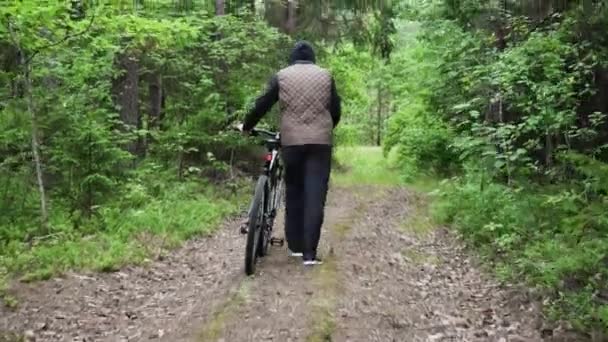 Een man loopt en rolt een fiets naast hem langs een bospad. Fiets defect, schade, pech. Zicht op de fietsers terug. Sport, recreatie en tijdverdrijf, gezondheidsvoordelen, fitness. Reis — Stockvideo