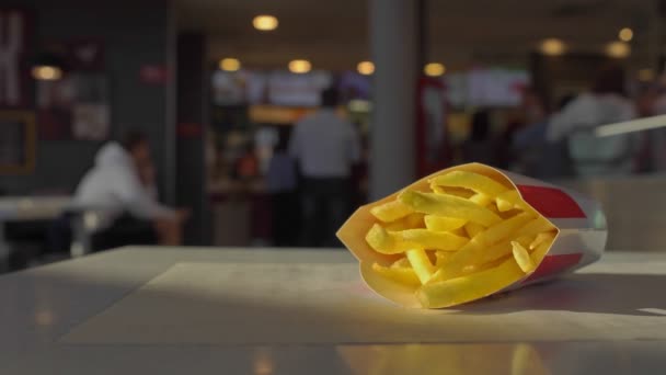 French fries potatoes lie on table inside cafe restaurant, mans hand takes and eats it. Fastfood. Unhealthy lifestyle, fried high calorie harmful food. Visitors in background make order near checkout — Wideo stockowe