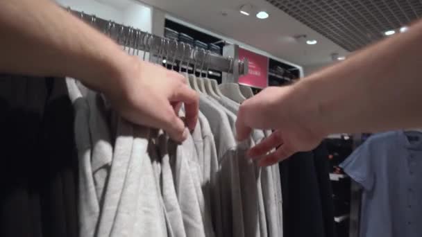 Egy férfi ruhát választ egy boltban, vásárol. Válogatás a pólók és kabátok a boltban, háttér, közelkép — Stock videók