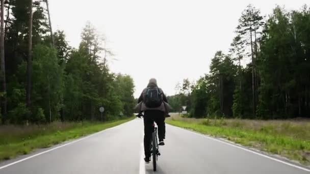 Guy with backpack rides cross-country mountain bike fast on an asphalt road through the green forest. View of the cyclists back. Sport, recreation and pastimes, health benefits, fitness. Trip journey — Stock Video
