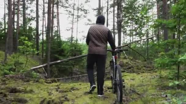 O tipo caminha e rola uma bicicleta ao lado dele, chegou a um beco sem saída na floresta, um obstáculo no caminho. Mau funcionamento da bicicleta, danos, avaria. Vista dos ciclistas de volta. Desporto, recreação. Viagem de viagem — Vídeo de Stock