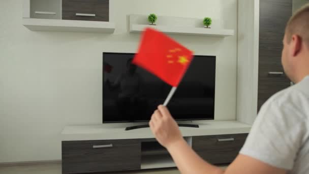 スポーツファンはテレビの前に家に座って、彼は中国の旗を振って、夏の冬のゲームイベントの競争で全国チームの勝利を祝います。男はワールドカップの放送を見ている。勝つ — ストック動画