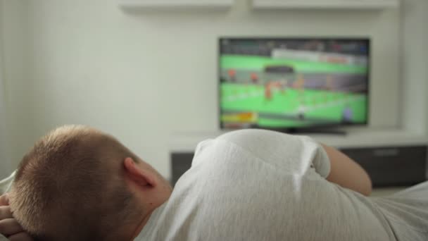 Мужской вентилятор лежит на диване в комнате дома и смотрит спортивное событие по телевизору. Летние Олимпийские игры. Лёгкая атлетика 2021 года. Корнеплод смотрит трансляцию на плазме в современном интерьере. Токио — стоковое видео