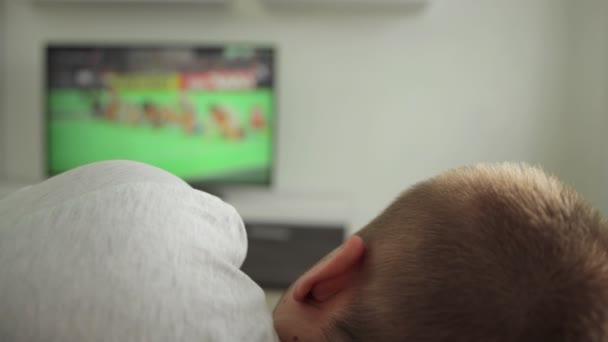 Man buff ligger på soffan i rummet hemma och tittar på sportevenemang på TV. Sommar olimpiska spel tävling. Friidrott kör 2021. Rooter fläkt klockor sänds på plasma i modern inredning. Från Tokyo — Stockvideo