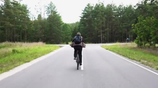Man with backpack rides cross-country mountain bike without pedaling on asphalt road through green forest. View of the cyclists back. Sport, recreation, health benefits, fitness. Trip journey — Stock Video
