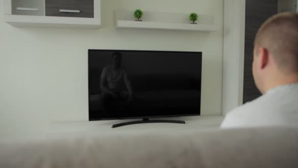 Killen sitter i hallen hemma och tittar på nyheter på cd-tv. Sport, film. Ett fan. Strömmande sändning. Fjärrkontroll i handen. Inomhusunderhållning på soffan i huset. Modern inredning lägenhet. Vad händer? — Stockvideo