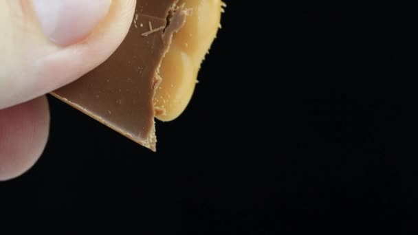 Un hombre rompe una barra de chocolate en sus manos de la que sale el relleno de turrón. Fondo negro, macro. Dulce postre, cámara lenta — Vídeo de stock