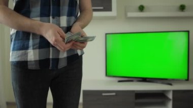 Adam evdeki televizyonun yanında elinde dolarları sayıyor. Yeşil ekran krom anahtarı. Bahis konsepti, bahse girerim. Kripto bozuk para piyasasında finansal ve yatırım başarısı. Modern iç daire odası.
