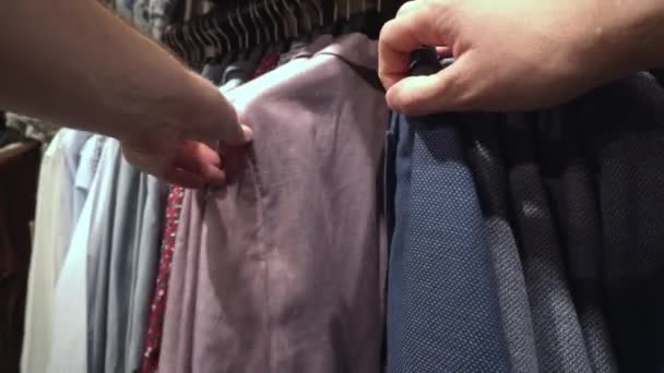 Egy férfi divatos ruhákat, kabátokat, ingeket és pólókat választ a boltban. Háttér, közelkép, vásárlások — Stock videók