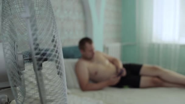 Ένας άντρας ξαπλώνει στον καναπέ στο σπίτι και κοιτάζει μέσα σε ένα smartphone. Το δωμάτιο ψύχεται το καλοκαίρι από έναν ανεμιστήρα στη θερμότητα για να δημιουργήσετε μια άνετη θερμοκρασία. Αντιγραφή χώρου για κείμενο — Αρχείο Βίντεο