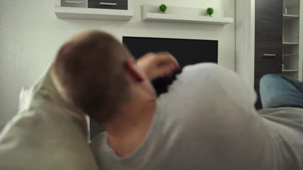 Der Mensch legt sich auf das Sofa, um zu Hause im Flur Nachrichten, Filme, Filme anzusehen. Fan beobachtet Sportereignis Spiel Wettbewerb. Guy sieht Streaming-Übertragung auf LCD in moderner Innenwohnung — Stockvideo