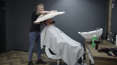 Berber salonu içinde. İnce berber müşterilerin yüzüne havlu atar. Erkek uzman, ziyaretçi kafasındaki kılları siler. Tımar, saç kesimi ve tıraş hizmeti. Pelerinli beyaz adam sandalyede oturuyor.