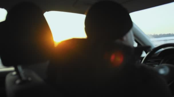 Een mannelijke chauffeur met jas en hoed rijdt 's winters in een auto op de snelweg. Een straal van de ondergaande zon schijnt door het raam, reis. Langzaam. — Stockvideo