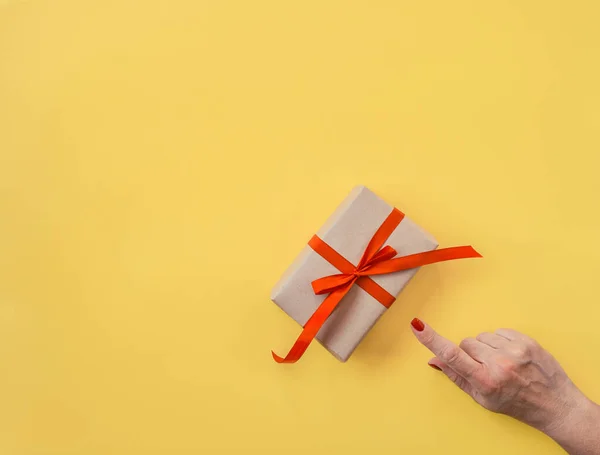 Woman hand points finger gift decorated with red ribbon, yellow background, copy space. Flat lay, hand and present box, top view. Valentine or love, spring holidays, Christmas and birthday concept.