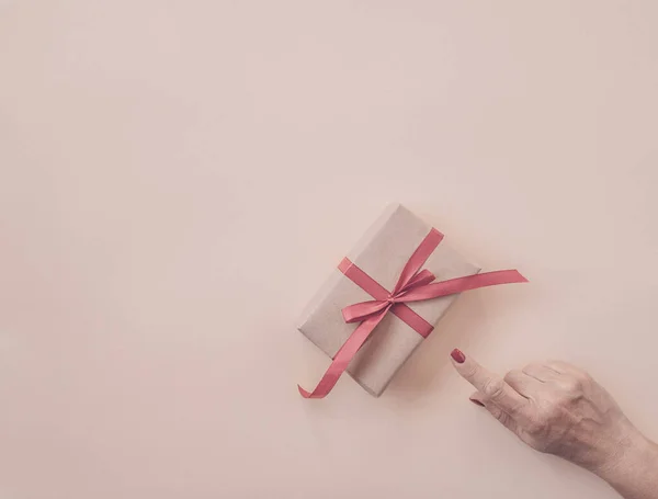 Woman hand points finger gift decorated with red ribbon, pink  background, copy space. Flat lay, hand and present box, top view. Valentine or love, spring holidays, Christmas and birthday concept.