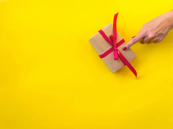 Woman hand touching finger, gift decorated with red ribbon, yellow background, copy space. Hand and present box, top view. Valentine or love, spring holidays, Christmas and birthday concept.