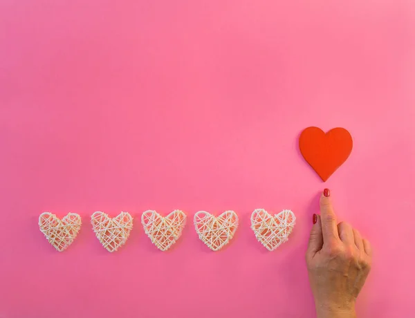 White wicker hearts line on pink background, copy space. Woman hand pointing finger red heart, top view. Valentine or love, spring holidays, Christmas and birthday concept.