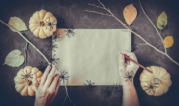 Halloween grunge black background, pumpkins, black spiders, dry branches, orange leaves decoration. Autumn composition frame, flat lay, vintage paper, woman hand with pencil, copy space.