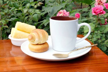 Beyaz kahve fincanı, hamur işleri ve bir gül bahçesindeki ahşap bir masada atıştırmalıklarla kahvaltı. Sıcak, doğal doğa manzarası yemek ve yaşam tarzı temaları için ideal.