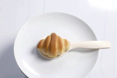 Seramik tabakta beyaz bir kaşığa yerleştirilmiş küçük bir kruvasan. Menüler, reklamlar ve stok fotoğrafçılığı için uygun minimum ve temiz gıda tasarımı, Top view