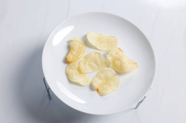 Beyaz bir tabağa yerleştirilmiş ince çıtır patates cipsi parlak stüdyo ışıklarıyla tabakta çıtır patates cipsi doku ve basit bir yemek sunumunu gösteriyor.