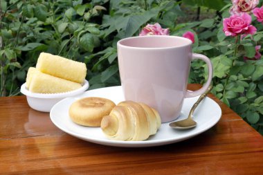 Pembe bir kahve fincanı, hamur işleri ve gül bahçesindeki ahşap bir masada atıştırmalıklarla kahvaltı. Sıcak, doğal doğa manzarası yemek ve yaşam tarzı temaları için ideal.