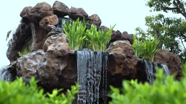 Verimli bir bahçede kayaların üzerinden akan yapay şelale. Yeşil bitkiler ve doğal ortam ile Bahçe Şelalesi doğa ve rahatlama temaları için huzurlu, ferahlatıcı bir açık hava sahnesi oluşturur