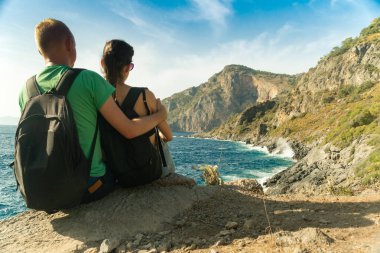 Sırt çantalı genç bir çift gün batımında kayalık sahilden denize bakarak kayaların üzerinde dinleniyor. Arka planda gezgin.