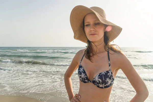 Bikinili rahat bir kadın tropikal kumsalın ve yaz tatilinin tadını çıkarıyor. Bronzlaşmış esmer, şapkalı, deniz kenarında yürüyüş yapan.