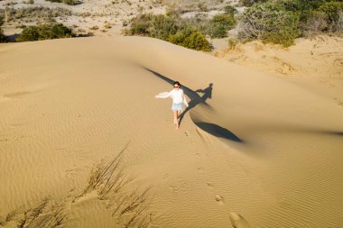 Çölde dalgalı kum tepelerinde duran kadın altın gün batımı ışığı ve rüzgarda.