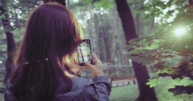 Cep telefonlu neşeli bir kadın parktaki orman ağacında günbatımının huzurlu bir fotoğrafını çekiyor. Güzel doğa günbatımı manzarası, akıllı telefonlu seyahat tatili fotoğrafı..